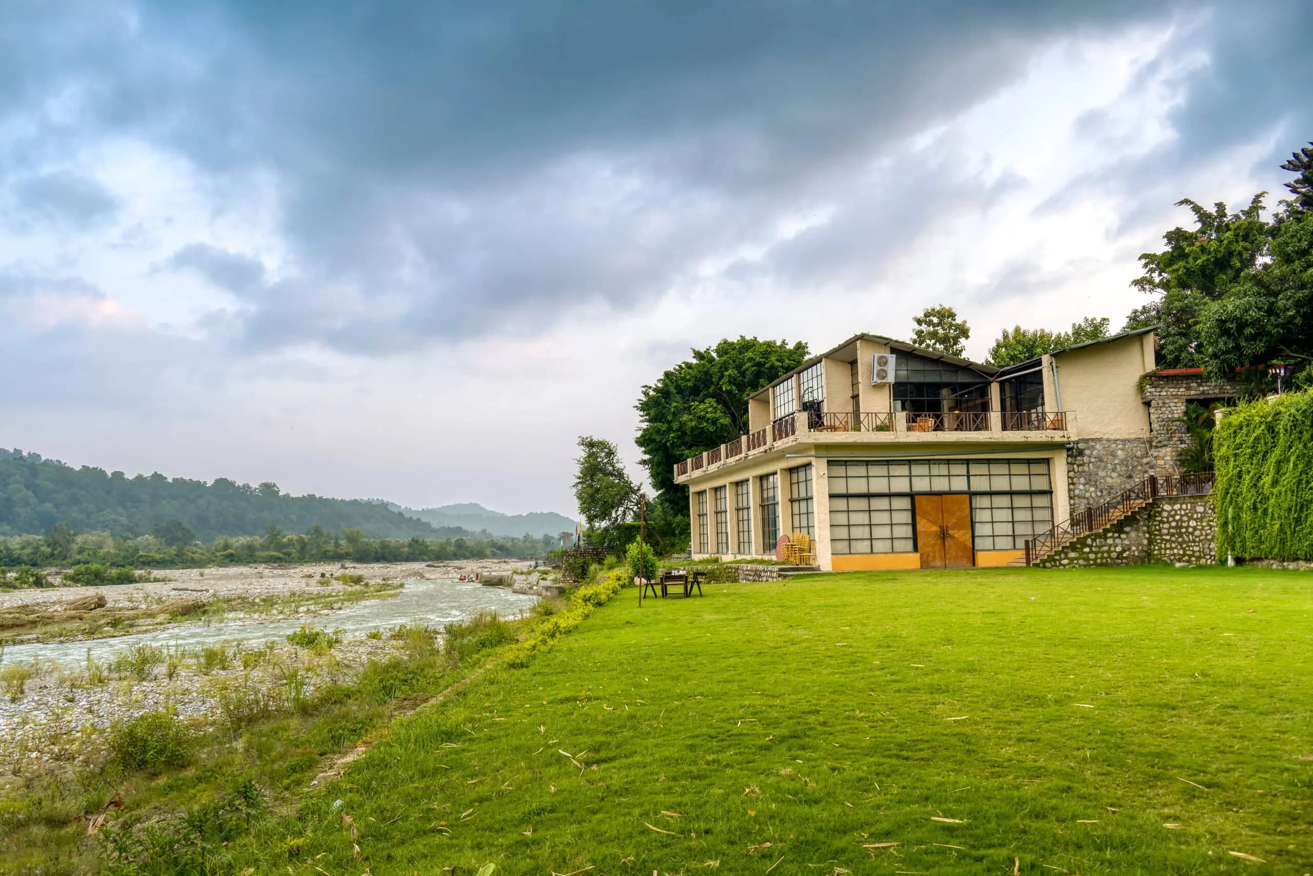 Scenic view of a river-facing resort property with modern architecture, a stone facade, and expansive lawn under a dramatic, cloudy sky.